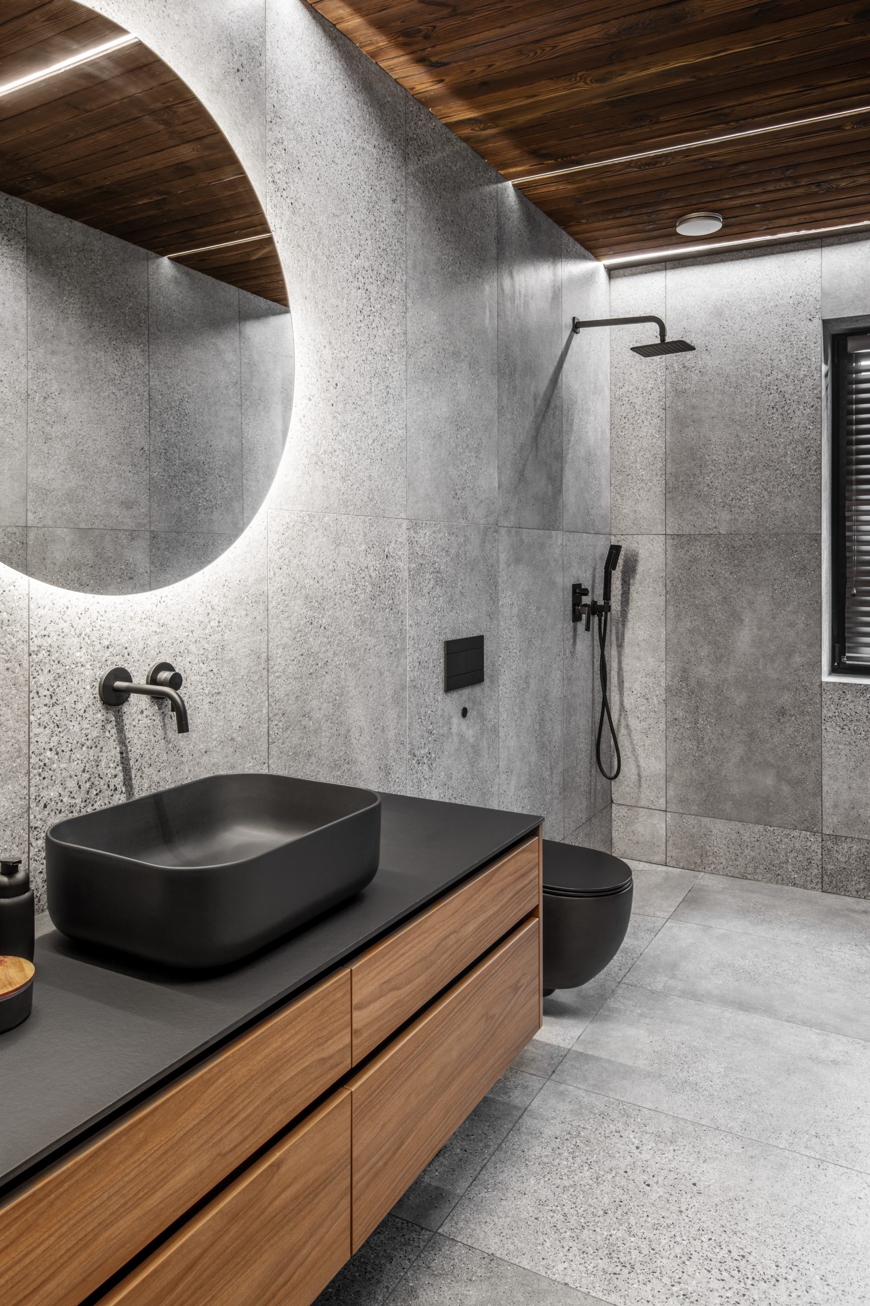 Modern minimalist bathroom interior with stone grey tiles, wooden furniture, black sink and toilet and round mirror. Spa bathroom concept